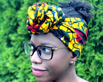 May include: A woman wearing a black shirt and glasses with a yellow, red, and green patterned head wrap.