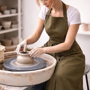 May include: A person working on a pottery wheel, shaping a clay pot. The person is wearing a white shirt and a green apron. The pottery wheel is white and blue. Shelves with pottery are in the background.