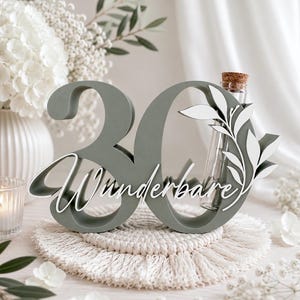 May include: A sage green wooden number 30 sign featuring the white cursive text Wunderbare and a white leaf cutout overlay. A clear glass test tube with a cork stopper is integrated into the right side of the zero, designed to hold small stems.