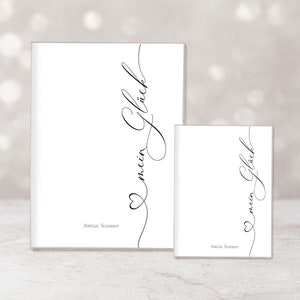 May include: Two white notebooks, one large and one small, with the words "mein Gl&uuml;ck" in elegant black script. The design includes a heart and the name "Amelie Schmidt" at the bottom. The background is a soft, blurred neutral color.