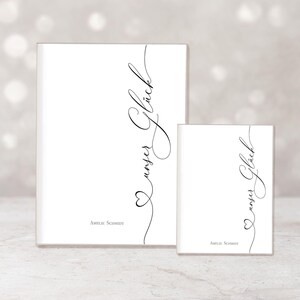 May include: Two white rectangular objects, one large and one small, with the words "unser Gl&uuml;ck" in elegant black script. A heart shape is incorporated into the script. The name "Amelie Schmidt" is printed at the bottom.