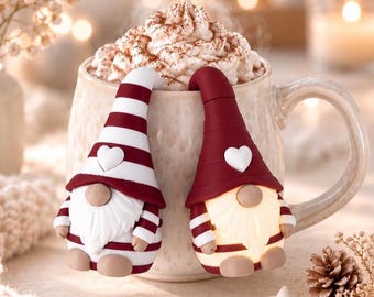 May include: Two decorative gnome ornaments perched on the rim of a ceramic mug filled with hot cocoa topped with whipped cream. One gnome wears a red and white striped hat, the other a solid red hat, both featuring a small white heart.