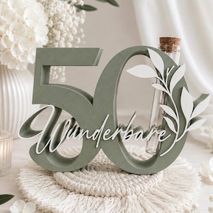May include: A sage green wooden number 50 sign featuring the white cursive text "Wunderbare" and a white leaf cutout design. A clear glass test tube with a cork stopper is integrated into the number zero, providing a space for small floral stems.