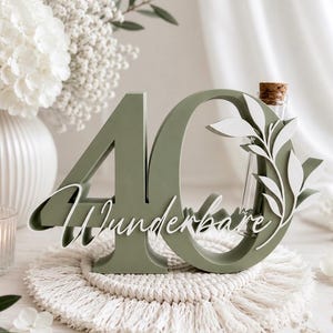 May include: A sage green wooden number 40 sign featuring the white cursive text Wunderbare and a white leaf cutout overlay. A clear glass test tube with a cork stopper is integrated into the zero, providing a space for a single stem or flower.
