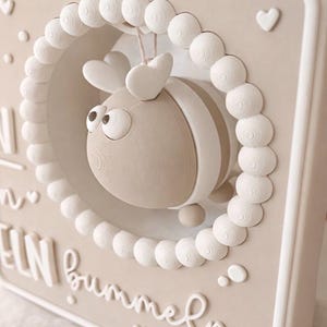 May include: A beige and white 3D wall art piece featuring a stylized bee inside a circular frame of white spheres. The bee has heart-shaped antennae and striped details. The background has text and heart accents.