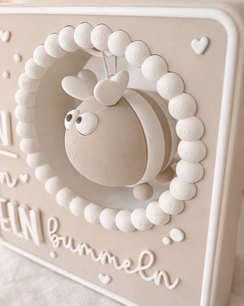 May include: A beige and white 3D wall art piece featuring a stylized bee inside a circular frame of white spheres. The bee has heart-shaped antennae and striped details. The background has text and heart accents.