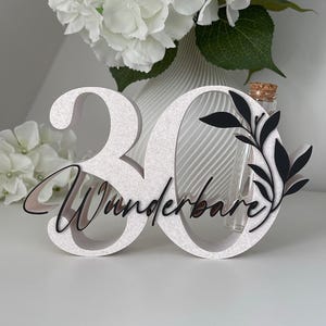 May include: A white number 30 sign with a textured finish, featuring the black script text "Wunderbare" and a black leaf graphic. A clear glass test tube with a cork stopper is integrated into the number zero, positioned upright.