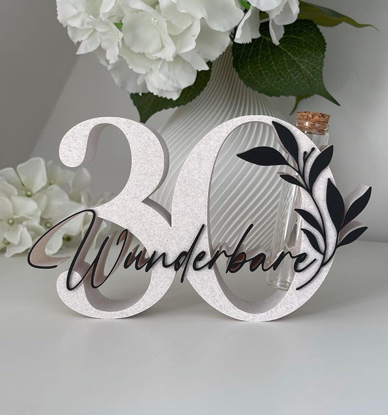 May include: A white number 30 sign with a textured finish, featuring the black script text "Wunderbare" and a black leaf graphic. A clear glass test tube with a cork stopper is integrated into the number zero, positioned upright.