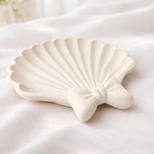May include: Off-white seashell-shaped decorative dish with a bow accent. The dish has a textured surface and a scalloped edge. The bow is centered at the base of the shell. The dish is on a white background.