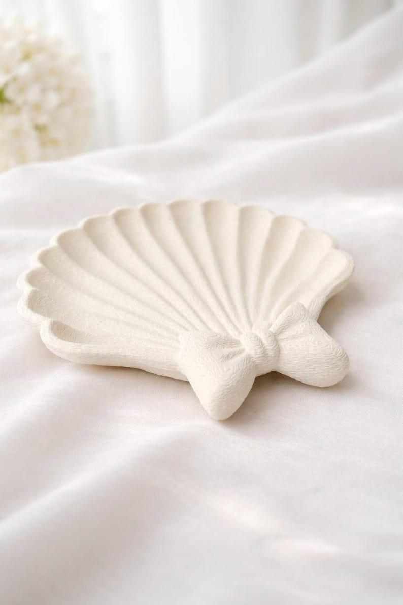 May include: Off-white seashell-shaped decorative dish with a bow accent. The dish has a textured surface and a scalloped edge. The bow is centered at the base of the shell. The dish is on a white background.