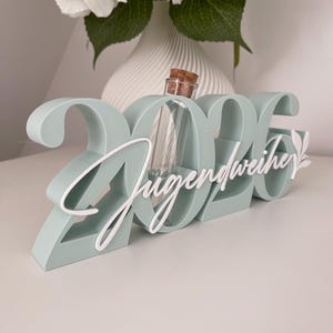 May include: A mint green decorative piece with the numbers "2026" and the word "Jugendweihe" in white script. A small glass bottle with a cork stopper is nestled within the numbers. A white floral arrangement is in the background.