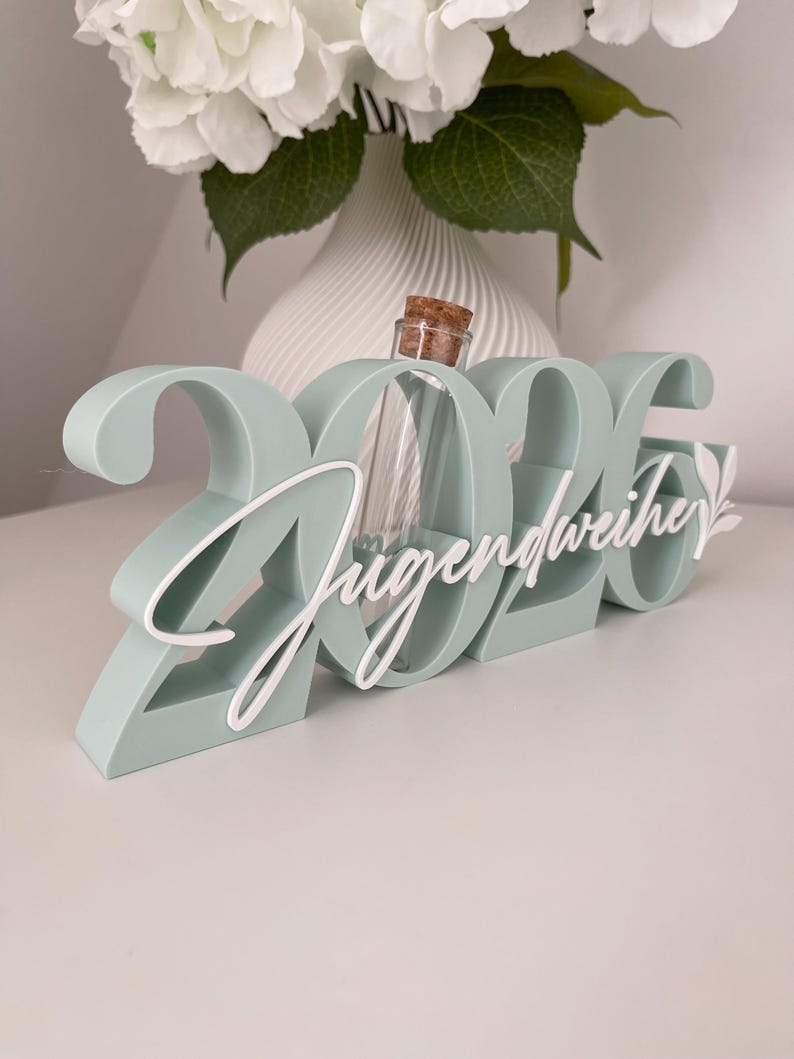 May include: A mint green decorative piece with the numbers "2026" and the word "Jugendweihe" in white script. A small glass bottle with a cork stopper is nestled within the numbers. A white floral arrangement is in the background.
