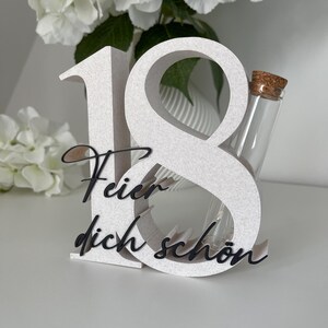 May include: A white, textured number 18 with the German phrase "Feier dich schon" in black script. A small glass vial with a cork stopper is nestled within the number 8. The background features white flowers.