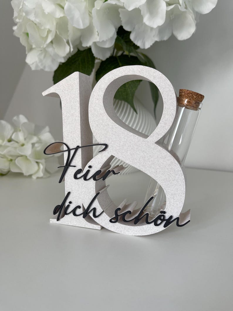 May include: A white, textured number 18 with the German phrase "Feier dich schon" in black script. A small glass vial with a cork stopper is nestled within the number 8. The background features white flowers.