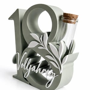 May include: A gray decorative piece in the shape of the number 18, with the word "Vollj&auml;hrig" in white script. A small glass bottle with a cork stopper is nestled within the design, along with white leaf accents.