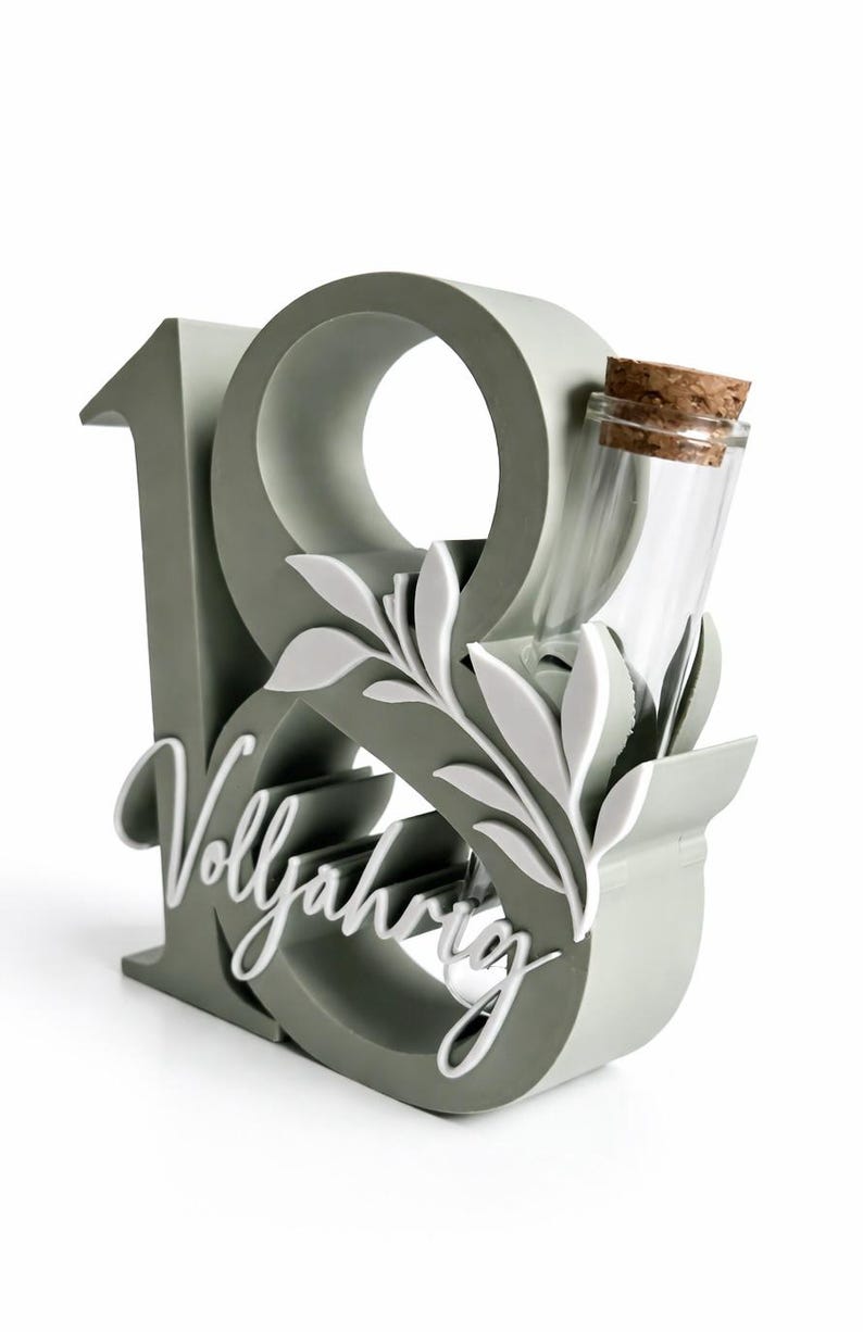 May include: A gray decorative piece in the shape of the number 18, with the word "Vollj&auml;hrig" in white script. A small glass bottle with a cork stopper is nestled within the design, along with white leaf accents.