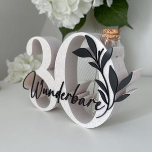 May include: A white number 30 display piece featuring the word Wunderbare in black script lettering. The number zero includes a black leaf design and a clear glass test tube vase with a cork stopper inserted into the top of the structure.