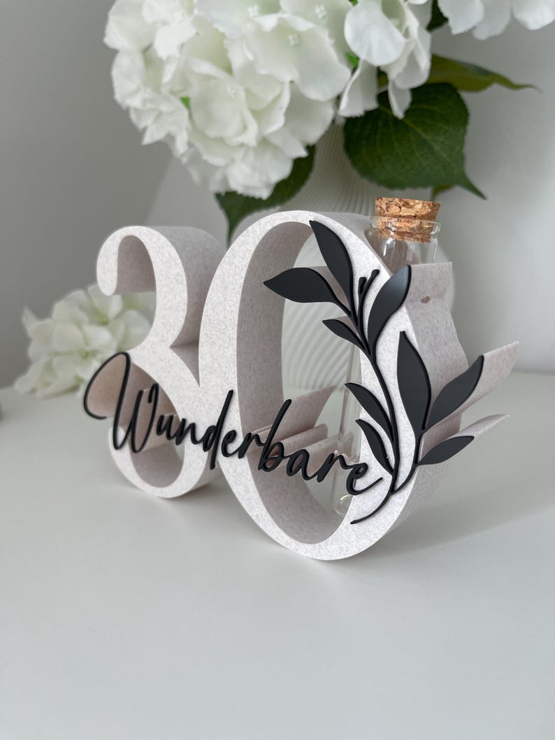 May include: A white number 30 display piece featuring the word Wunderbare in black script lettering. The number zero includes a black leaf design and a clear glass test tube vase with a cork stopper inserted into the top of the structure.