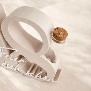 May include: A light beige decorative object with the word "Q" and the words "Feier dich sch&ouml;n" in white script. A small clear glass bottle with a cork stopper is nestled within the "Q".
