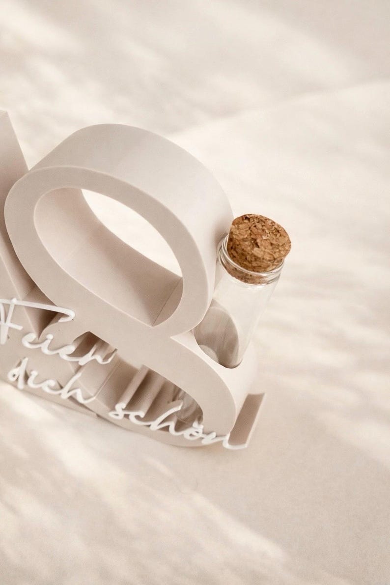 May include: A light beige decorative object with the word "Q" and the words "Feier dich sch&ouml;n" in white script. A small clear glass bottle with a cork stopper is nestled within the "Q".