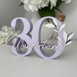 May include: A light purple 3D number 30 sign featuring the white cursive text "Wunderbare" and a white leaf cutout. A clear glass test tube with a cork stopper is integrated into the structure of the number zero, providing a space for small items.