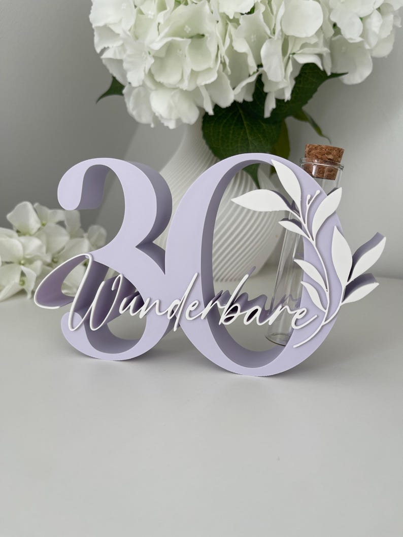 May include: A light purple 3D number 30 sign featuring the white cursive text "Wunderbare" and a white leaf cutout. A clear glass test tube with a cork stopper is integrated into the structure of the number zero, providing a space for small items.