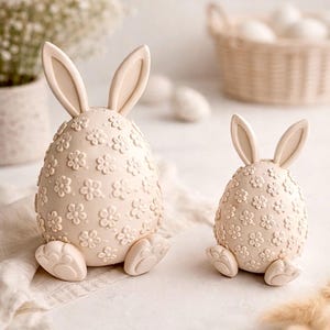 Könnte beinhalten: Zwei cremefarbene Osterei-Hasenfiguren. Die eiförmigen Körper sind mit kleinen, geprägten Blumen verziert. Die Hasen haben aufrechte Ohren und sitzen mit ausgestreckten Füßen. Im Hintergrund ist ein Korb mit Eiern zu sehen.