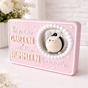 May include: A rectangular pink decorative sign featuring the German text "Bin im GARTEN mit den HUMMELN bummeln" in white and beige lettering. A small, round, black and beige bumblebee figure hangs inside a white circular frame.