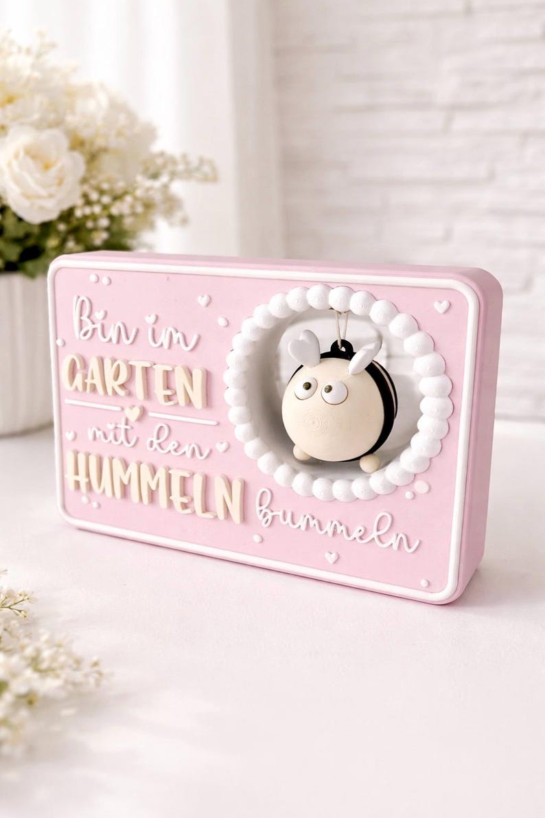 May include: A rectangular pink decorative sign featuring the German text "Bin im GARTEN mit den HUMMELN bummeln" in white and beige lettering. A small, round, black and beige bumblebee figure hangs inside a white circular frame.
