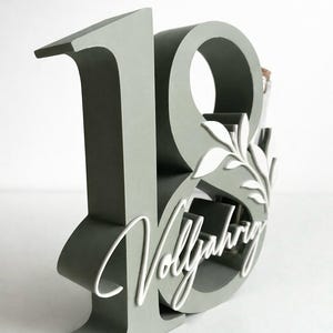 May include: A gray wooden number 18 with the word "Vollj&auml;hrig" in white script. The number has a decorative leaf design and a space for a small bottle. A unique gift for an 18th birthday.