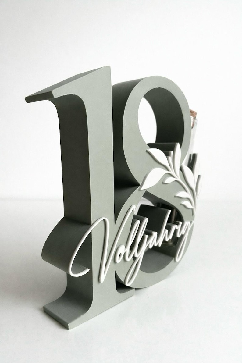 May include: A gray wooden number 18 with the word "Vollj&auml;hrig" in white script. The number has a decorative leaf design and a space for a small bottle. A unique gift for an 18th birthday.