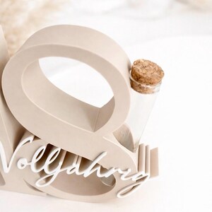 May include: A beige wooden display piece with the word "Vollj&auml;hrig" in white script. It features a large letter "O" and holds a small glass vial with a cork stopper, perfect for a small flower or keepsake.