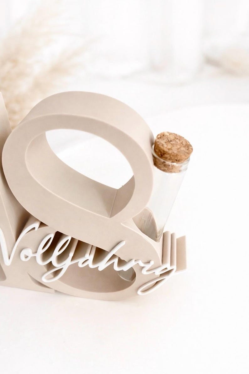 May include: A beige wooden display piece with the word "Vollj&auml;hrig" in white script. It features a large letter "O" and holds a small glass vial with a cork stopper, perfect for a small flower or keepsake.