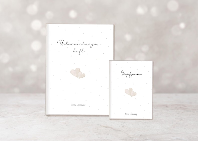May include: Two white notebooks with the text "Untersuchungsheft" and "Impfpass" in a delicate script. Each notebook features a watercolor illustration of two hearts and the name "Nina Lehmann".