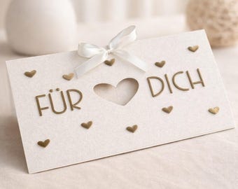 Puede incluir: Una tarjeta de felicitación crema con un recorte en forma de corazón y las palabras alemanas "FÜR DICH" en letras doradas. La tarjeta está adornada con pequeños adornos de corazón dorado y un lazo de cinta blanca. Un artículo decorativo para ocasiones especiales.