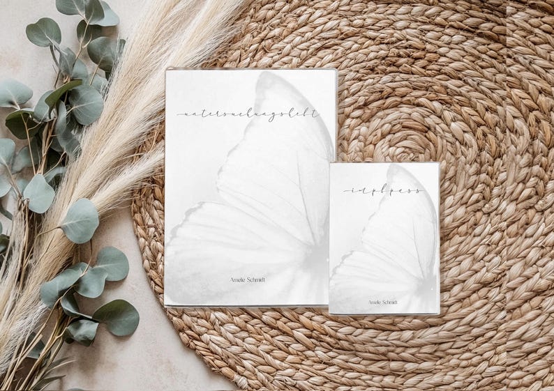 May include: Two white art prints with butterfly wing designs and handwritten text. The larger print reads "maternelangbeft" and the smaller one reads "-eapbess". Both prints are on a woven mat with eucalyptus and pampas grass.
