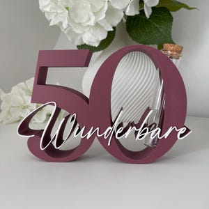 May include: A dark maroon wooden number 50 cutout featuring the white cursive text Wunderbare layered across the front. A clear glass test tube with a cork stopper is positioned vertically within the opening of the number zero.
