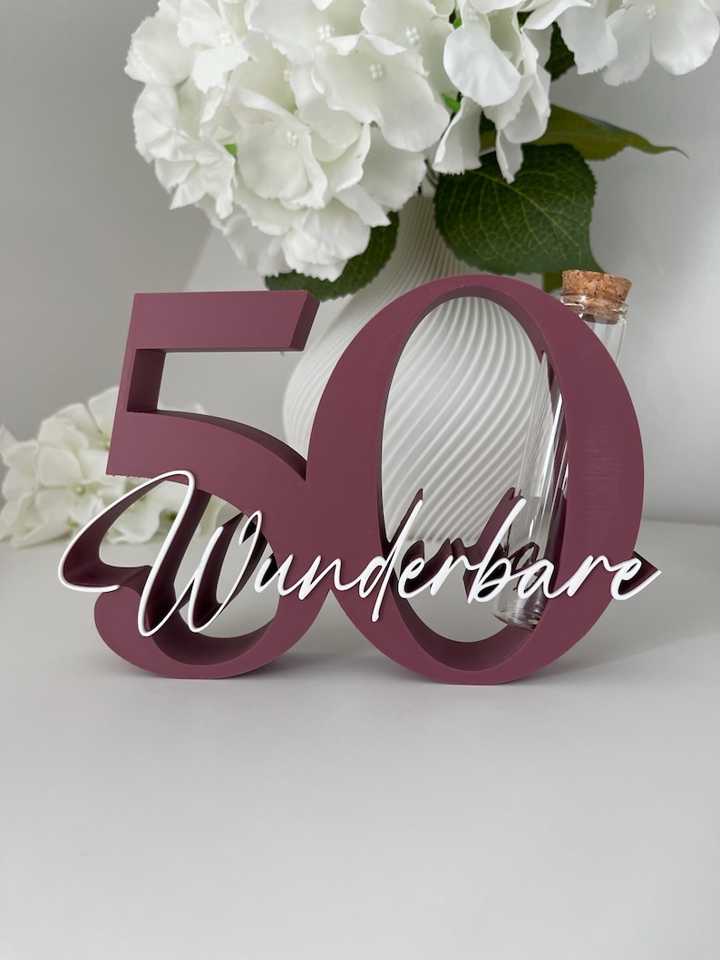 May include: A dark maroon wooden number 50 cutout featuring the white cursive text Wunderbare layered across the front. A clear glass test tube with a cork stopper is positioned vertically within the opening of the number zero.
