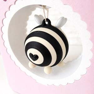 May include: A decorative ornament with a black and white striped pattern and a heart detail. The ornament has small white wings and is suspended within a pink, scalloped-edged frame. The design is whimsical and charming, suitable for gifting.