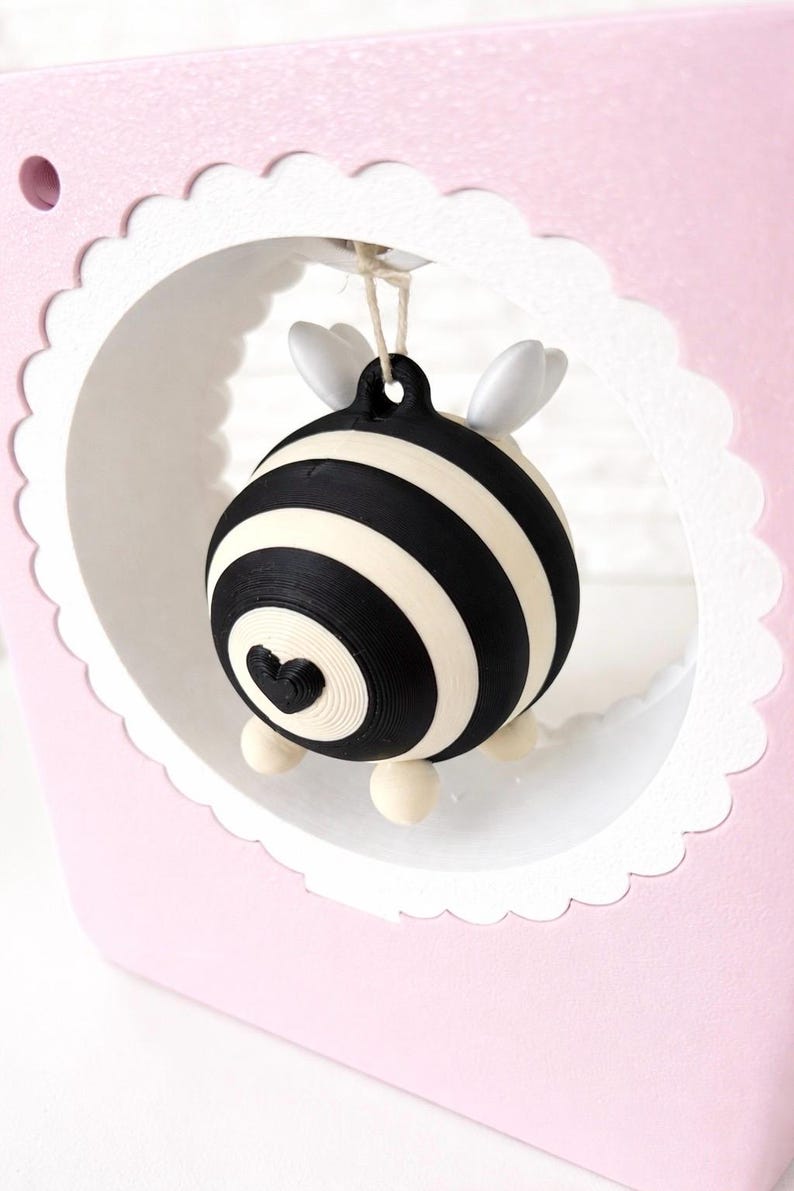 May include: A decorative ornament with a black and white striped pattern and a heart detail. The ornament has small white wings and is suspended within a pink, scalloped-edged frame. The design is whimsical and charming, suitable for gifting.