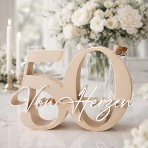 May include: A beige wooden number 50 decoration featuring the white cursive text Von Herzen layered across the front. A small clear glass bottle with a cork stopper is tucked into the opening of the number zero, creating a decorative display piece.