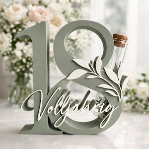 May include: A sage green wooden '18' with the word 'Vollj&auml;hrig' in white script, accented with white leaf details. A small glass vial with a cork stopper is nestled within the design, suitable for a milestone celebration.