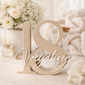 May include: A beige wooden number 18 with the word "Vollj&auml;hrig" in white script, likely for a birthday celebration. A small glass bottle with a cork stopper is visible. The background includes white flowers and a soft, neutral-toned setting.