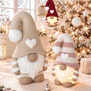 Könnte beinhalten: Zwei große Wichtelfiguren mit hohen Mützen, weißen Bärten und Herzapplikationen, eine in Beige und eine in Rosa, sowie ein winziger passender Wichtel. Ein kleiner Kreis zeigt einen ähnlichen Wichtel mit dunkelroter Mütze.