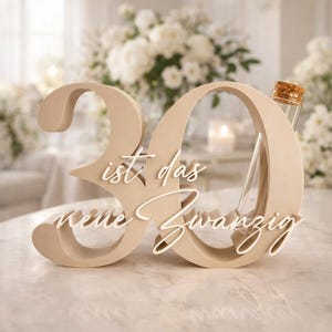 May include: A beige wooden cutout of the number 30 with white cursive text reading "ist das neue Zwanzig" layered across the front. A clear glass test tube with a cork stopper is inserted into the right side of the number zero.