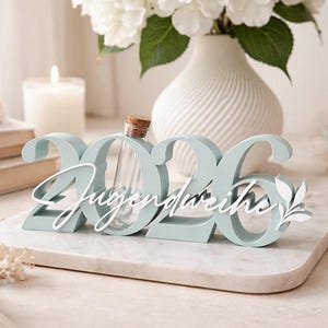 May include: A light blue decorative piece with the numbers "2026" and the word "Jugendweihe" in white script. A small glass vial with a cork stopper sits in the center. White flowers in a vase are in the background.