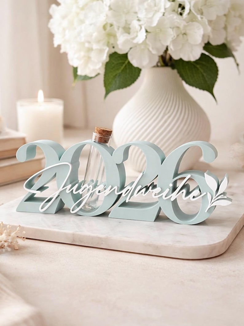 May include: A light blue decorative piece with the numbers "2026" and the word "Jugendweihe" in white script. A small glass vial with a cork stopper sits in the center. White flowers in a vase are in the background.