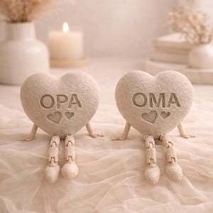 May include: Two heart-shaped figurines with articulated legs and arms. The beige hearts have the words "OPA" and "OMA" engraved, each with two heart symbols. The figurines are on a light-colored, textured surface.