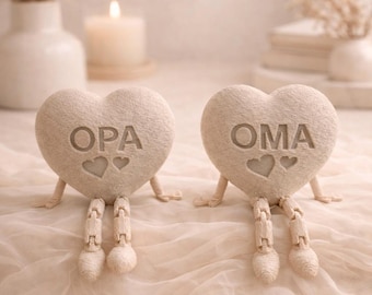 Op de afbeelding: Twee hartvormige beeldjes met beweegbare benen en armen. De beige harten hebben de woorden "OPA" en "OMA" gegraveerd, elk met twee hartsymbolen. De beeldjes staan op een lichtgekleurd, getextureerd oppervlak.