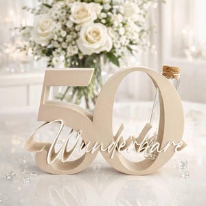 May include: A beige wooden cutout of the number 50 with the white cursive script word Wunderbare layered across the front. A small clear glass bottle with a cork stopper is tucked into the opening of the zero, standing upright.
